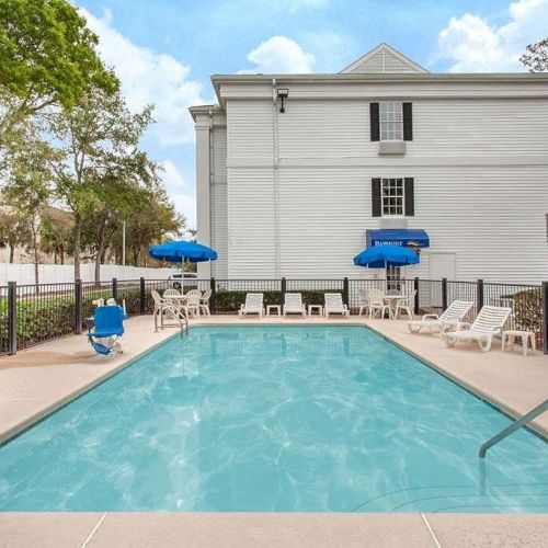 This image shows a rectangular outdoor swimming pool next to a white building with blue umbrellas, lounge chairs, and a pool lift.