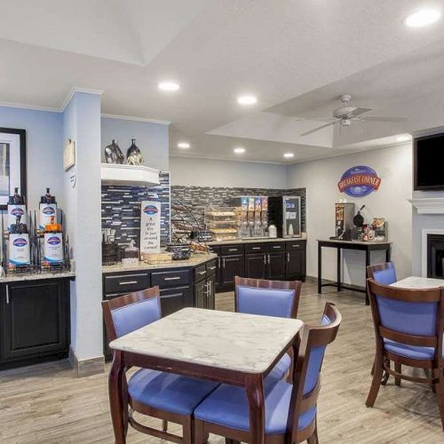 The image shows a dining area with tables and chairs, a coffee station, a microwave, a TV, and a breakfast area equipped with various food items, ending the sentence.