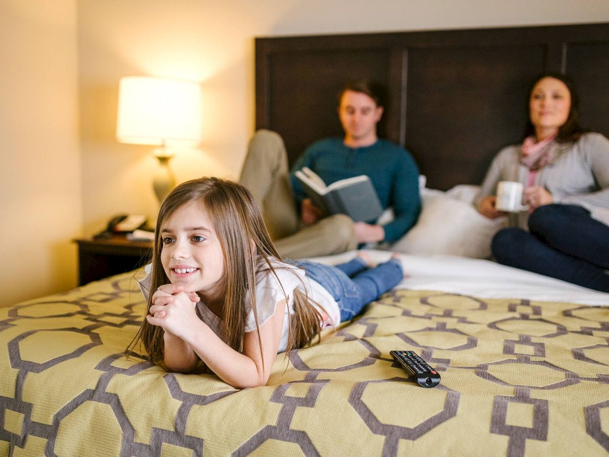 A young girl lies on a bed watching TV, with a remote beside her, while two adults sit behind her, one reading a book and the other holding a mug.