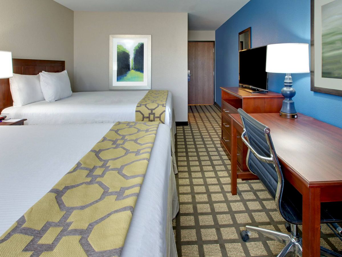 This image shows a hotel room with two beds, a desk with a chair, a flat-screen TV, and lamps on both the desk and beside the beds.