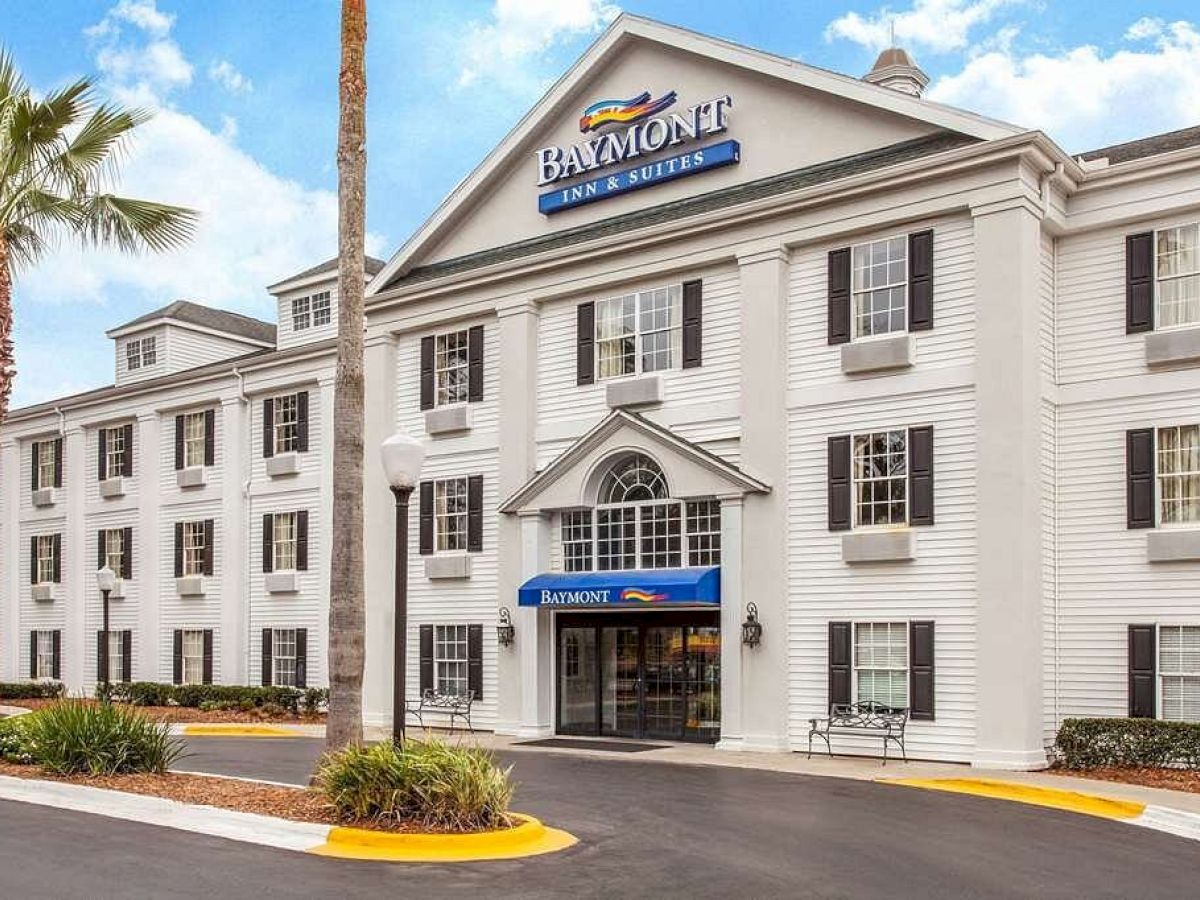 The image shows the exterior of a Baymont Inn & Suites hotel with three stories, white facade, black shutters, and a landscaped entrance.