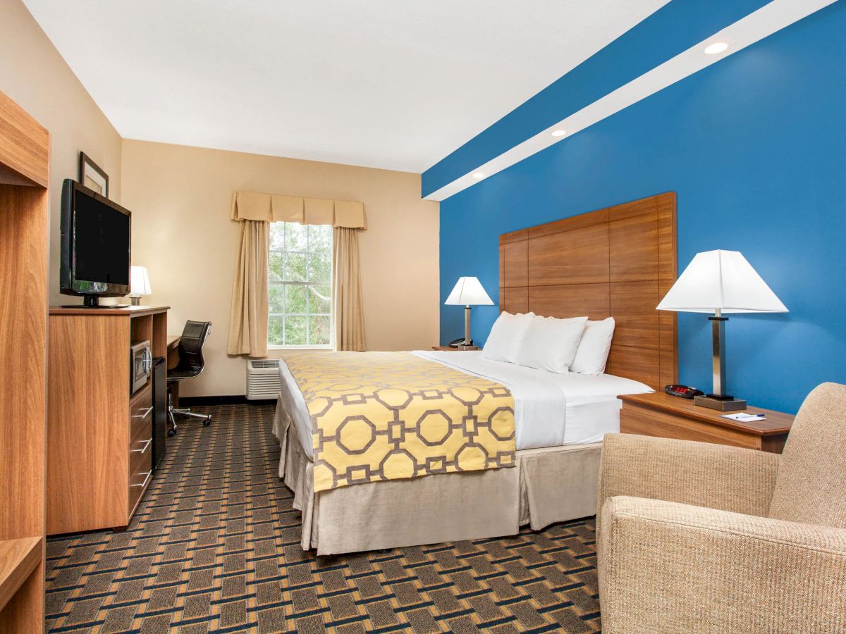 A hotel room with a bed, blue accent wall, desk with a TV, armchair, lamps, window with drapes, and a patterned carpet.