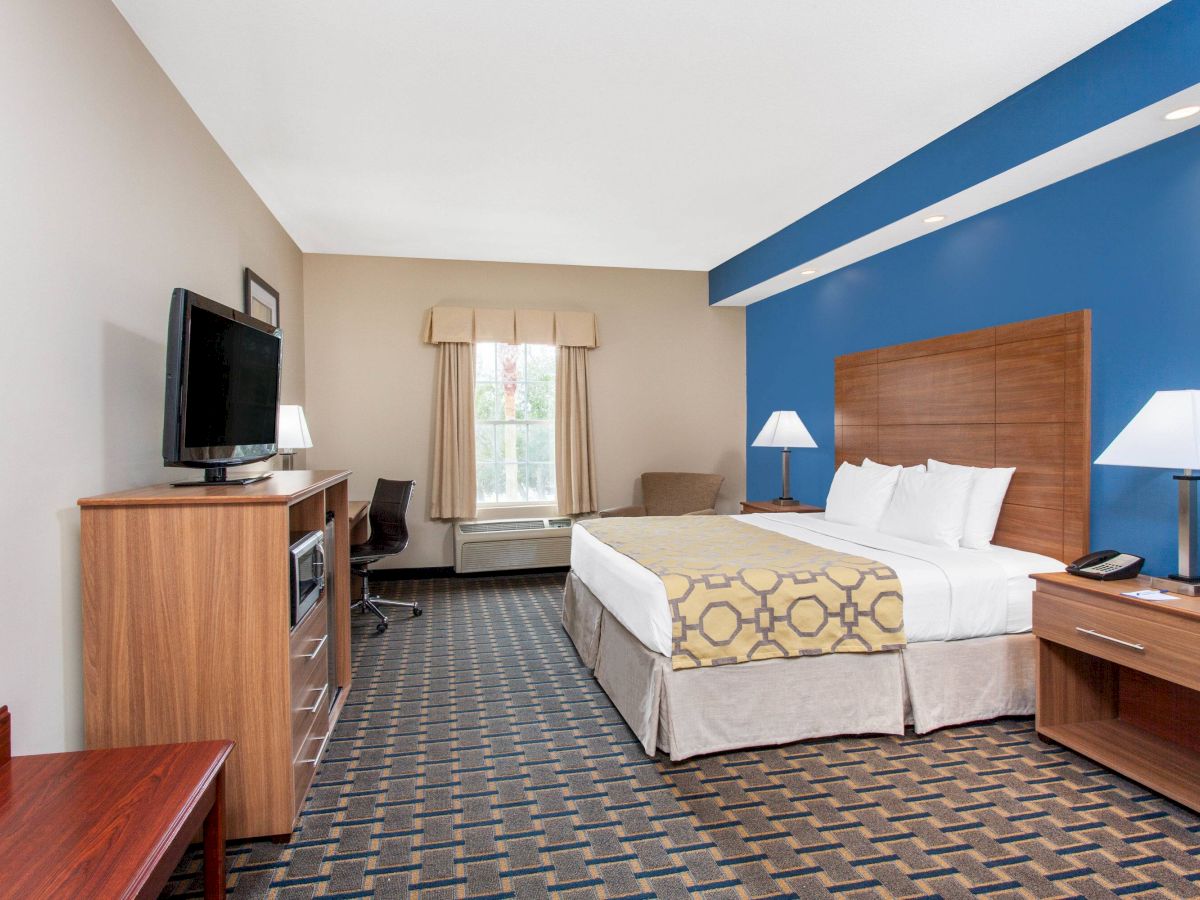 A hotel room with a king-size bed, two bedside tables, a flat-screen TV, a desk, a chair, a window with curtains, and a blue accent wall.