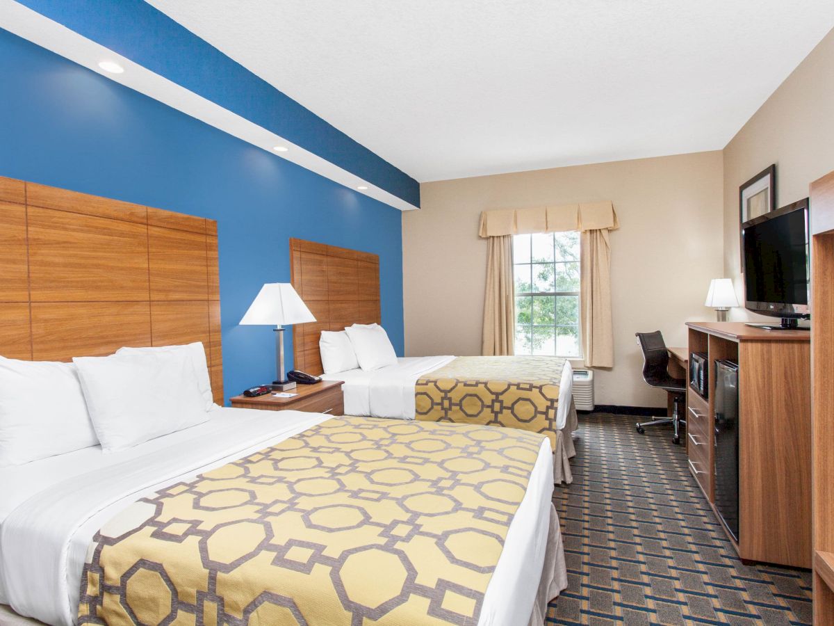 A hotel room features two beds, a blue accent wall, lamps, a TV, desk, chair, and a window with curtains, creating a cozy and functional space.