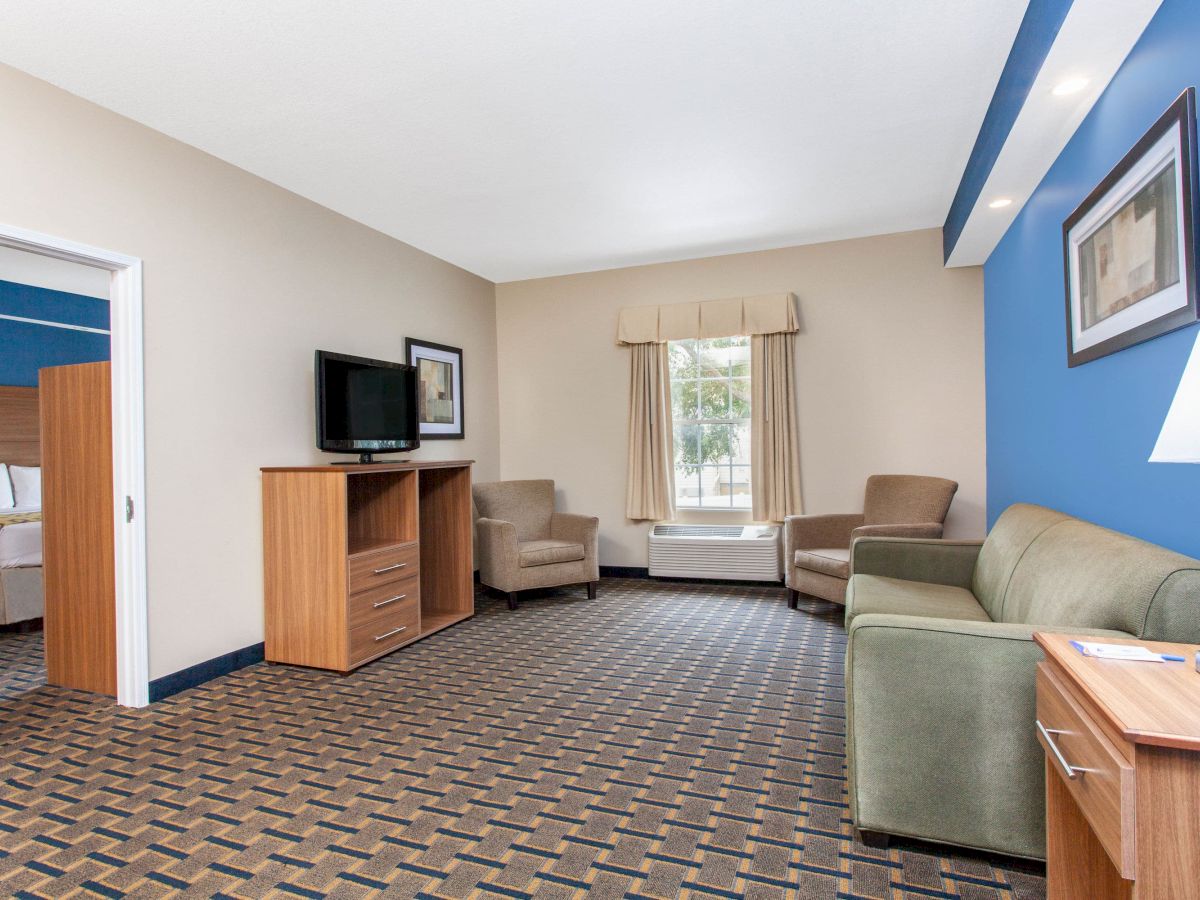 A hotel room features a sitting area with a green sofa, two chairs, a TV, and a view of a second bedroom through an open doorway.