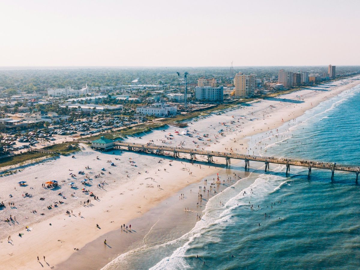 An aerial view of a bustling beach with people sunbathing and swimming, a pier extending into the ocean, and a cityscape in the background with various buildings.