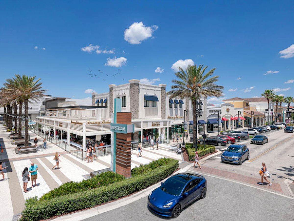 An outdoor shopping center with palm trees, shops, cars, and people walking around on a sunny day, featuring a sign that reads 