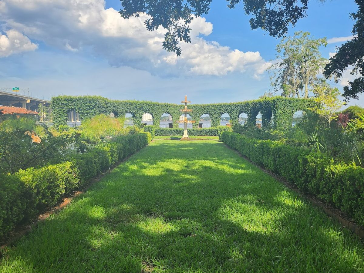 The image shows a garden path lined with trimmed hedges, leading to an archway covered in greenery under a bright blue sky with scattered clouds.