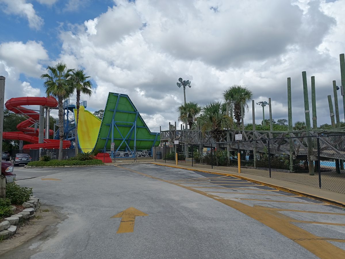 The image shows a water park with colorful slides, a large green and yellow water ride structure, palm trees, and a path leading through.