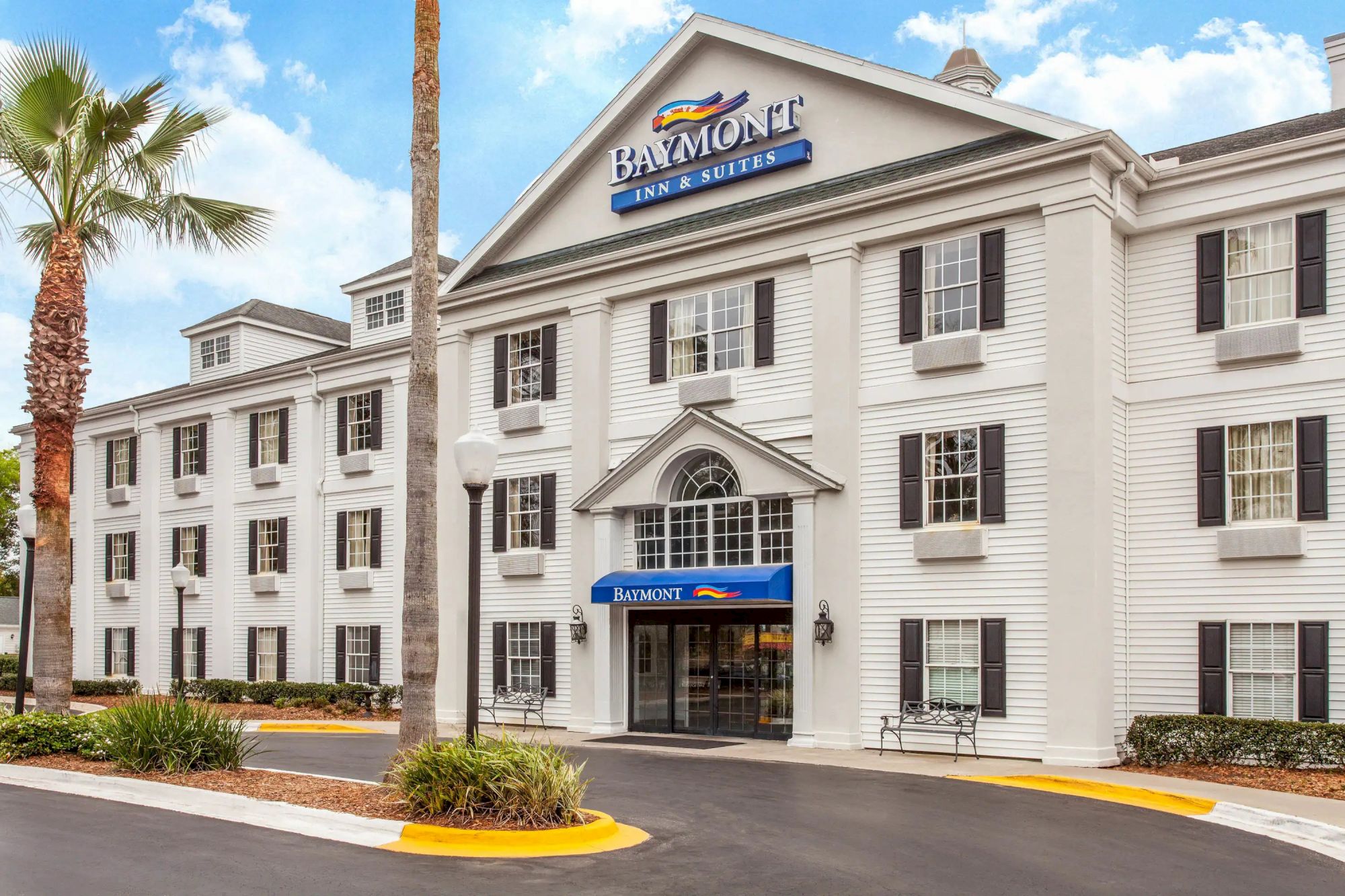 This image shows the exterior of a Baymont Inn & Suites hotel with a light-colored facade, multiple windows, and a landscaped area with palm trees.