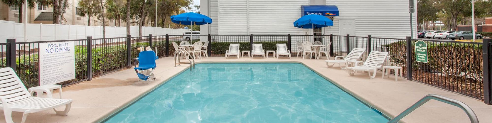 Outdoor pool with lounge chairs, blue umbrellas, and a white building in the background; surrounded by greenery and blue sky with some clouds.