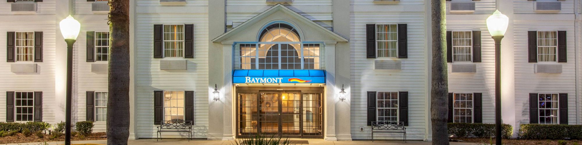 The image shows the exterior of a Baymont Inn & Suites hotel at dusk, with illuminated lamps and a well-maintained landscape.