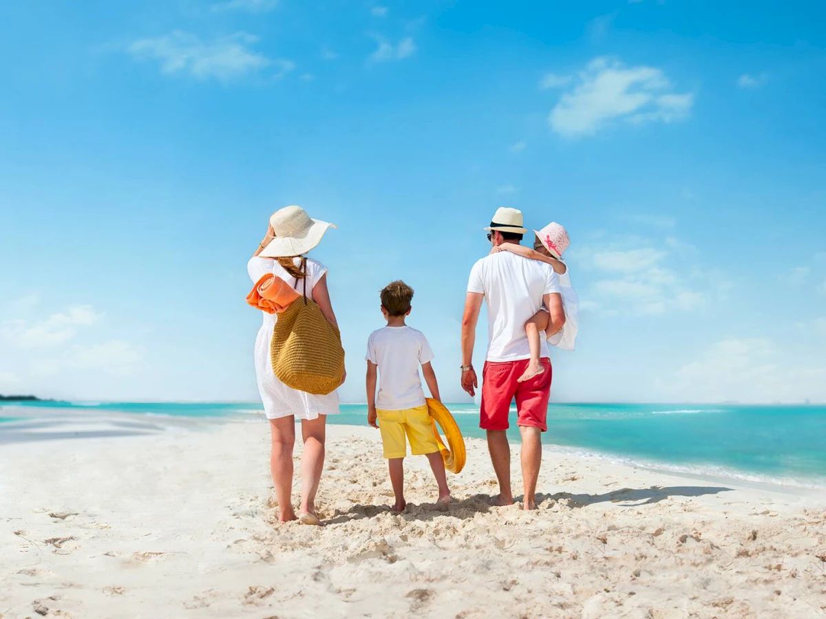 A family of four, two adults and two children, is walking on a sandy beach with clear blue skies and calm sea water in the background.