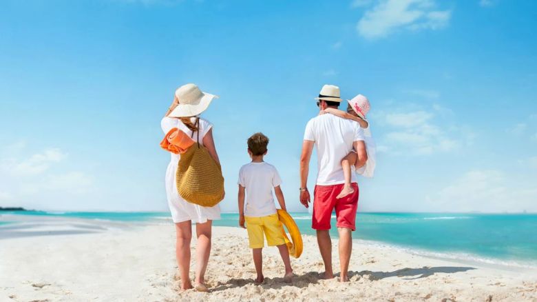 A family of four, two adults and two children, is walking on a sandy beach with clear blue skies and calm sea water in the background.