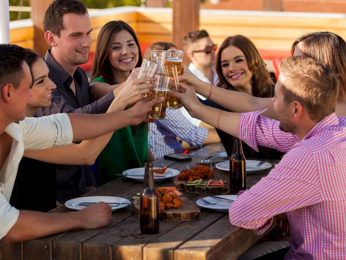 A group of people sitting around a wooden table, clinking glasses and smiling, with plates of food and drinks in front of them.