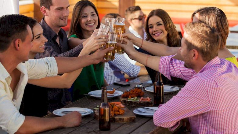 A group of people sitting around a wooden table, clinking glasses and smiling, with plates of food and drinks in front of them.