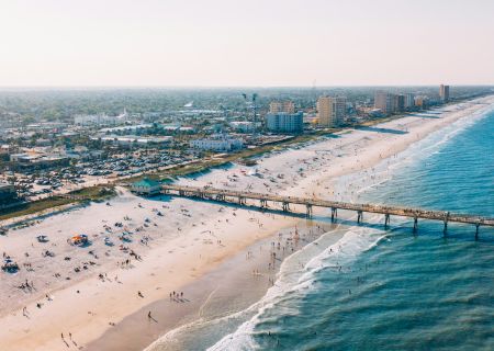 Aerial view of a beach with many people, a pier stretching into the ocean, and a coastal city in the background under a clear sky.