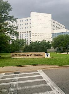 This image shows the exterior of the Mayo Building and Hospital with vehicles parked in front and a cloudy sky overhead, ending the sentence.