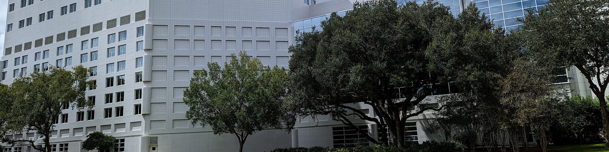 The image shows a large, modern building with multiple stories, adjacent trees, a grassy area, and a gravel path under a partly cloudy sky.