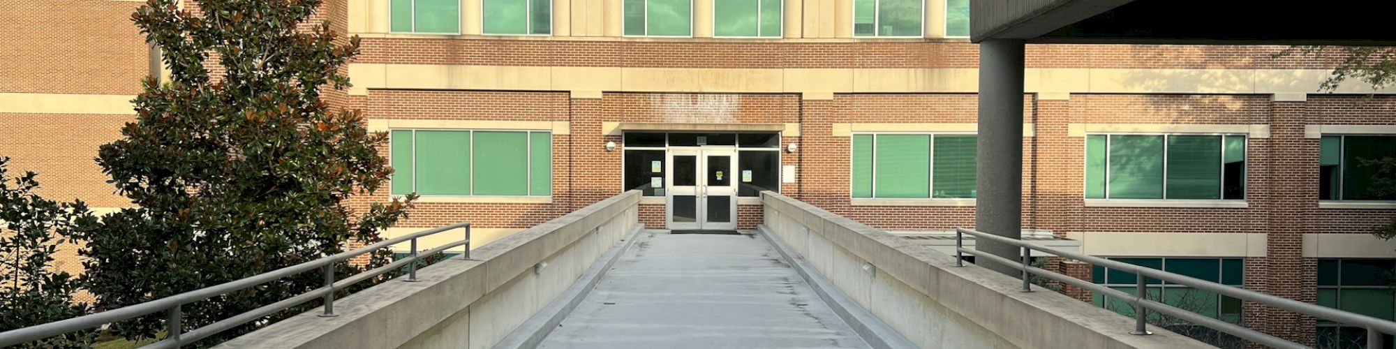 A walkway leads to the entrance of a multi-story brick building with windows, framed by another structure on the right and a tree on the left.