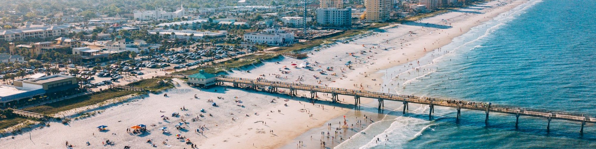 An aerial view of a coastal city with a sandy beach, a pier extending into the ocean, and buildings alongside the shore, on a sunny day.