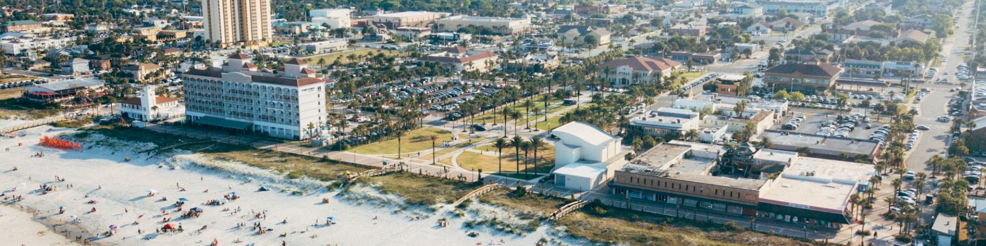 Aerial view of a coastal city with a crowded beach, hotels, and buildings in the background on a sunny day.