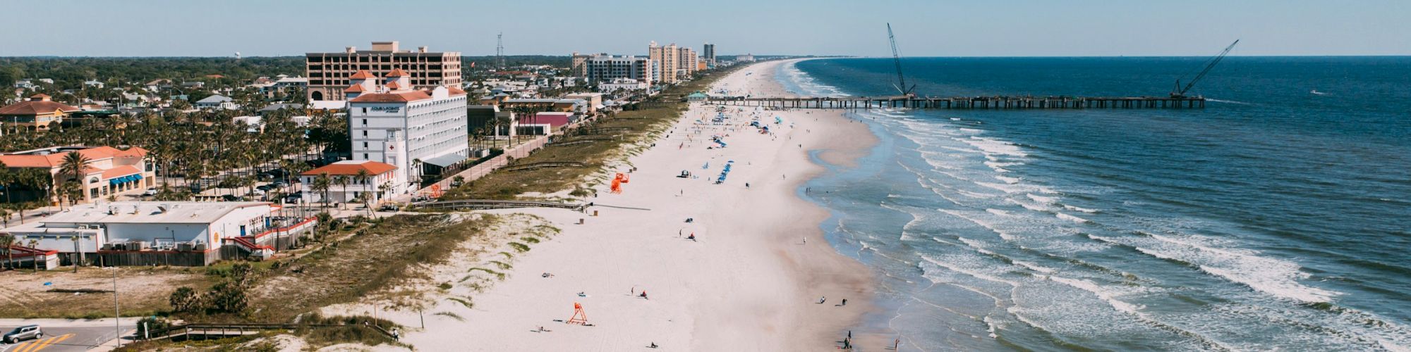 A shoreline view with a sandy beach, a few people, buildings in the distance, and a pier extending into the ocean under a clear sky.