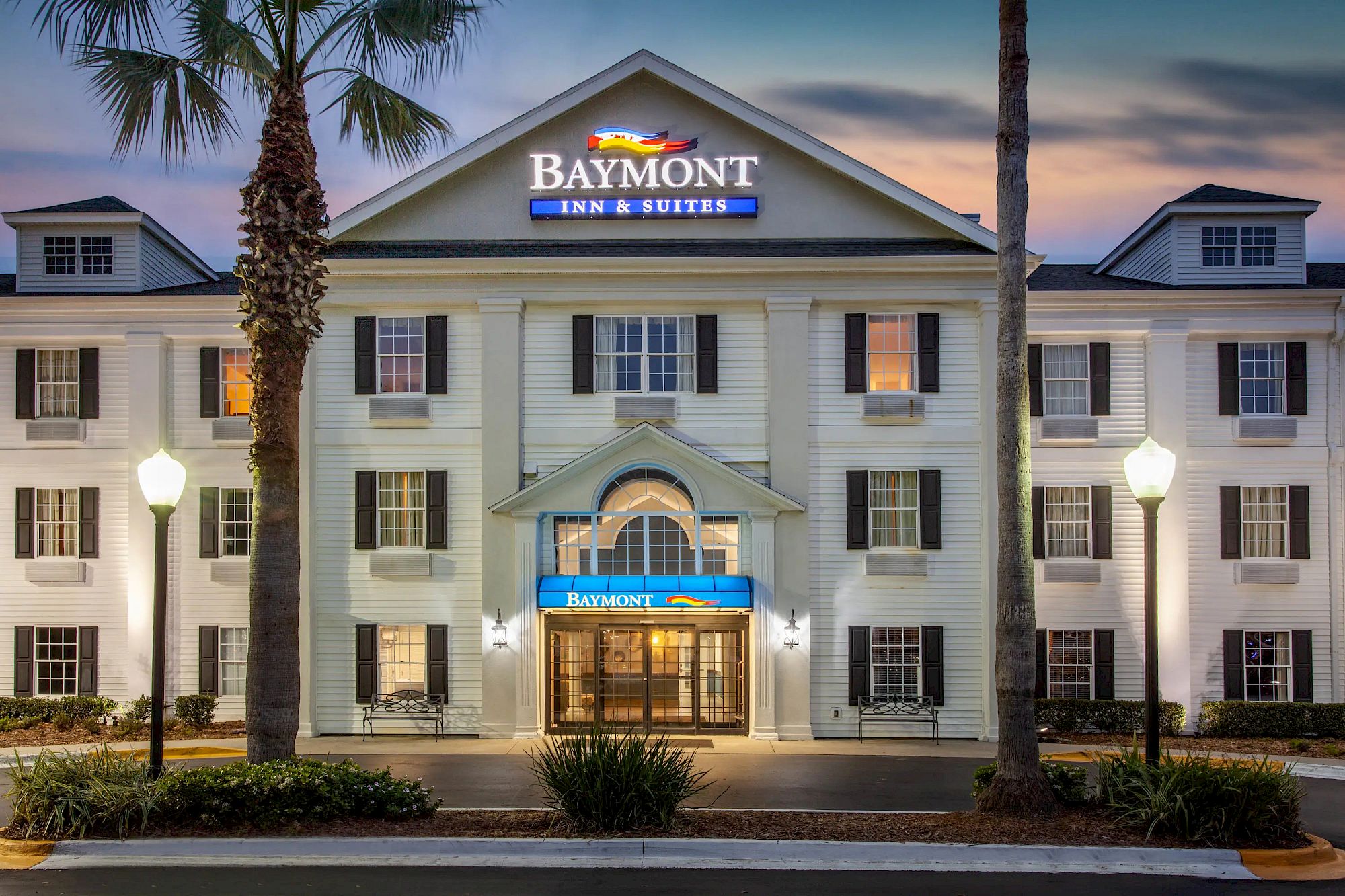 The image shows the exterior of a Baymont Inn & Suites hotel at dusk, with illuminated signs, palm trees, and street lights adorning the entrance.