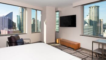 The image shows a modern hotel room with large windows offering a cityscape view, a bed, a seating area, a TV, and minimalistic furniture.