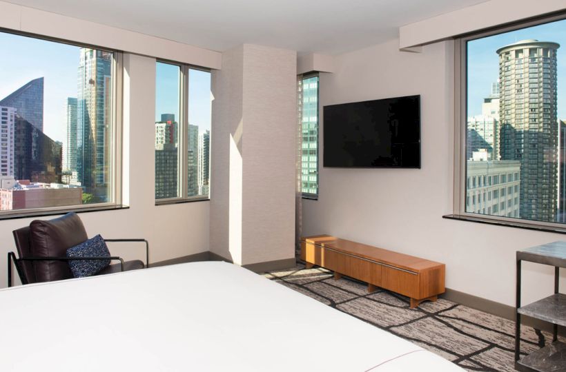 The image shows a modern hotel room with large windows offering a cityscape view, a bed, a seating area, a TV, and minimalistic furniture.
