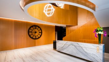 This image shows a modern reception area with a marble desk, wooden wall paneling, a large clock, and decorative lighting fixtures.