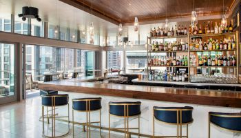The image shows a modern bar with a wooden counter, bar stools, and a fully stocked liquor shelf in a bright, open space with large windows.