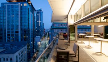 An outdoor terrace with tables and chairs overlooks a cityscape with tall buildings and a lit street, viewed during dusk.