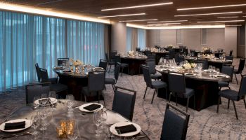 The image shows a modern dining room setup with round tables, black chairs, and elegant table settings, ready for an event in a softly lit room.
