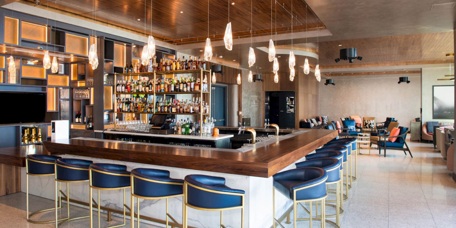 A modern bar with a U-shaped counter, blue bar stools, hanging lights, well-stocked shelves of bottles, and a lounge area with seating.
