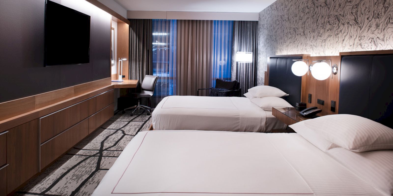 A modern hotel room with two double beds, a wall-mounted TV, a work desk, and soft lighting. The window has curtains partially drawn, enhancing coziness.