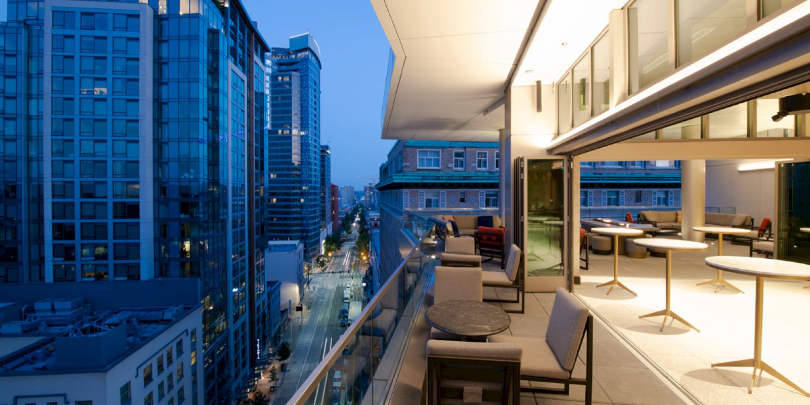 The image shows a modern balcony with seating areas overlooking a cityscape with high-rise buildings at dusk.