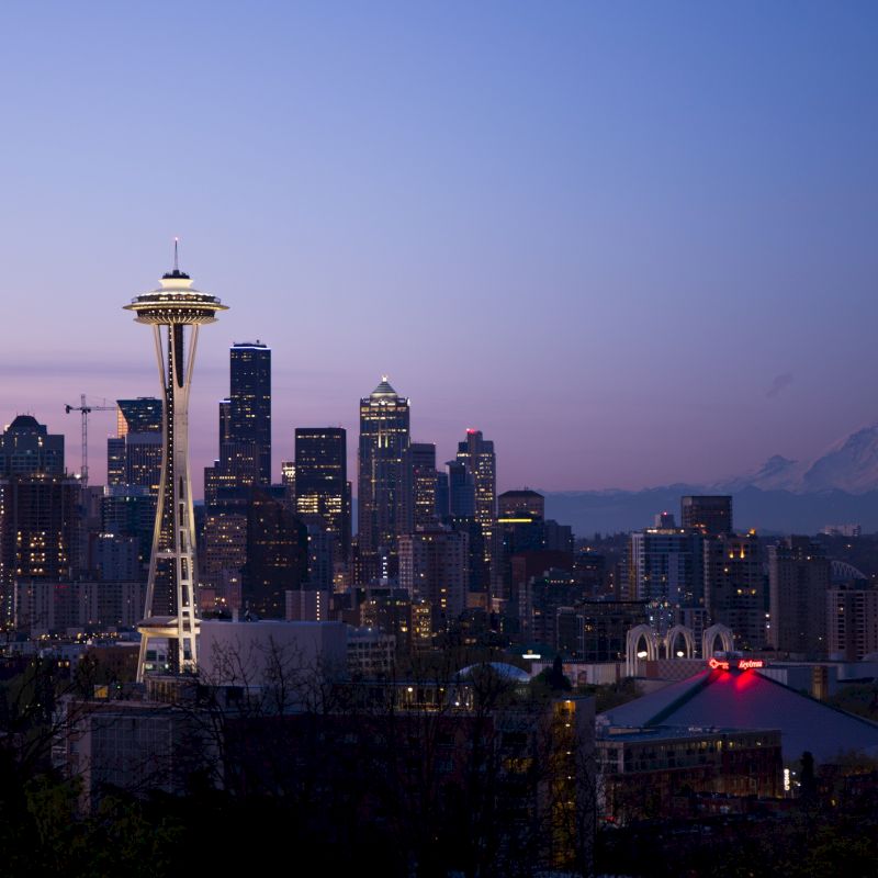 A cityscape at dusk with a prominent tower in the foreground, and a mountain visible in the background, under a purple and blue sky.