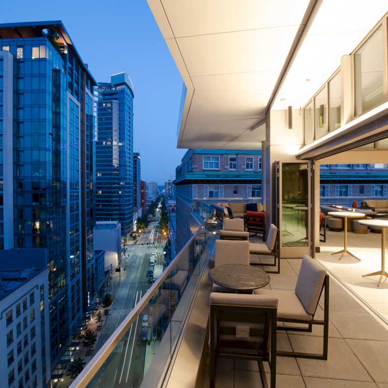 The image shows a modern balcony with seating overlooking a city street at dusk, with tall buildings on either side of the street.