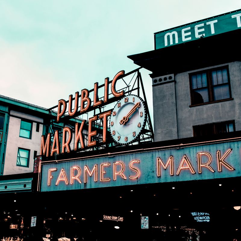 A famous public market sign with a clock and neon lights, reading "Farmers Market," against a backdrop of buildings.