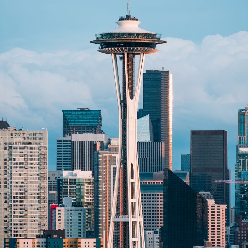 The image shows the Space Needle with surrounding buildings and skyscrapers, set against a partly cloudy sky in Seattle.