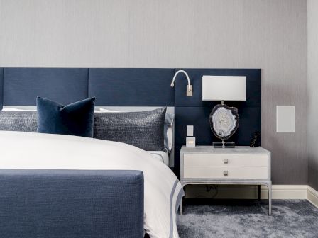 This image shows a modern bedroom with a blue headboard, white bedding, blue pillows, a nightstand with a lamp, and a decorative object.