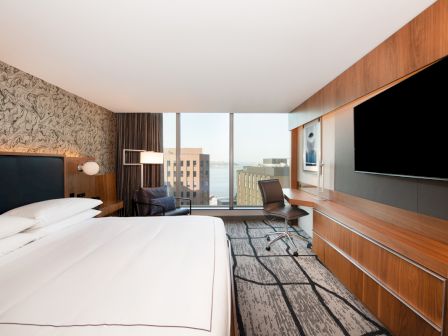A modern hotel room features a large bed, a wall-mounted TV, a desk with a chair, and a window with a city and water view ending the sentence.