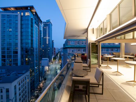 A modern balcony with a dining area overlooks a city street lined with tall buildings at twilight, featuring a sleek and open design.