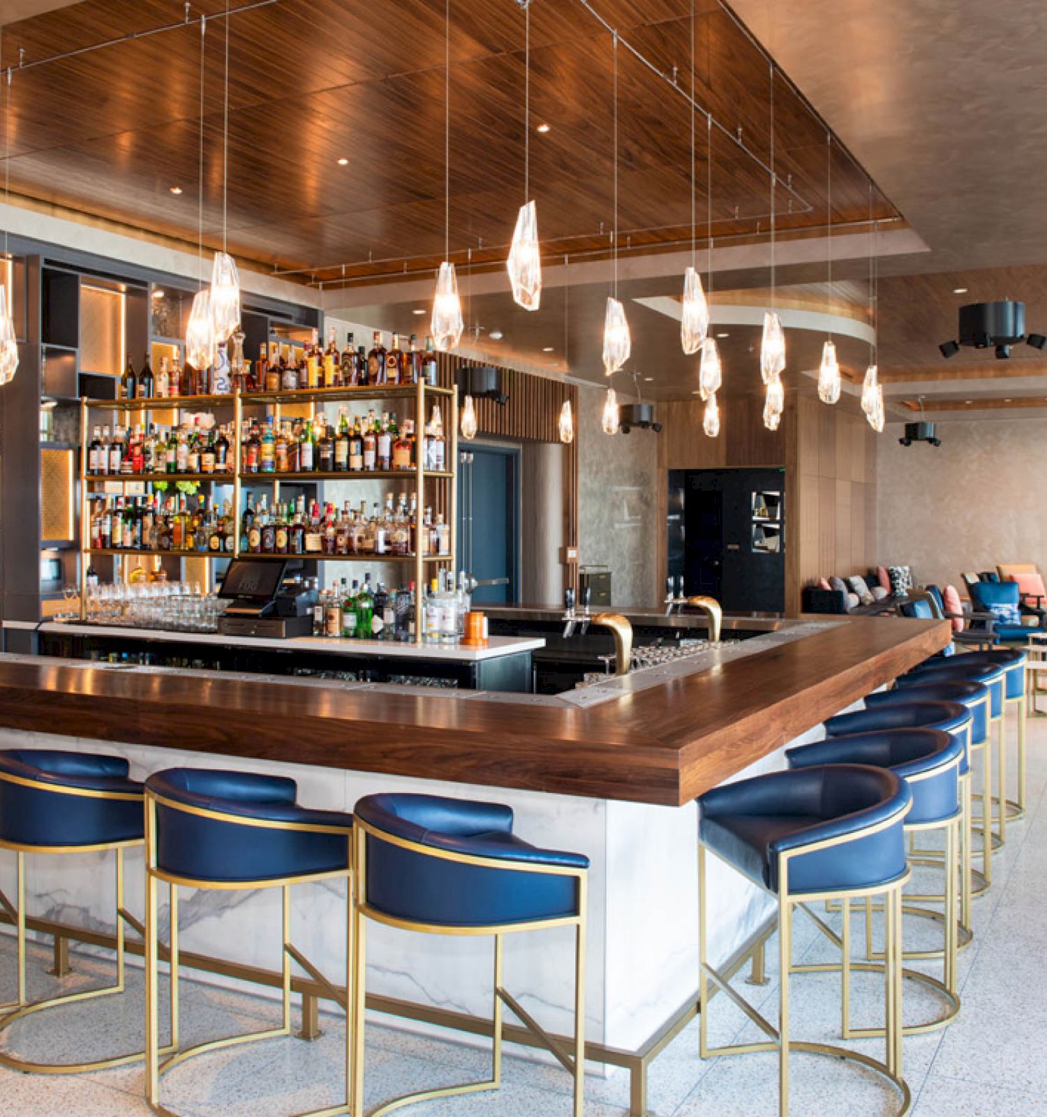 This image shows a modern bar with a wooden counter, blue bar stools, hanging lights, and a well-stocked glass shelving unit of various drinks.