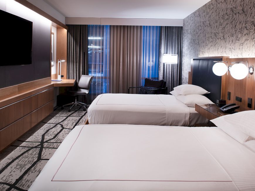 The image shows a modern hotel room with two beds, a TV, a desk with a chair, and minimalist decor, featuring neutral tones and a patterned carpet.