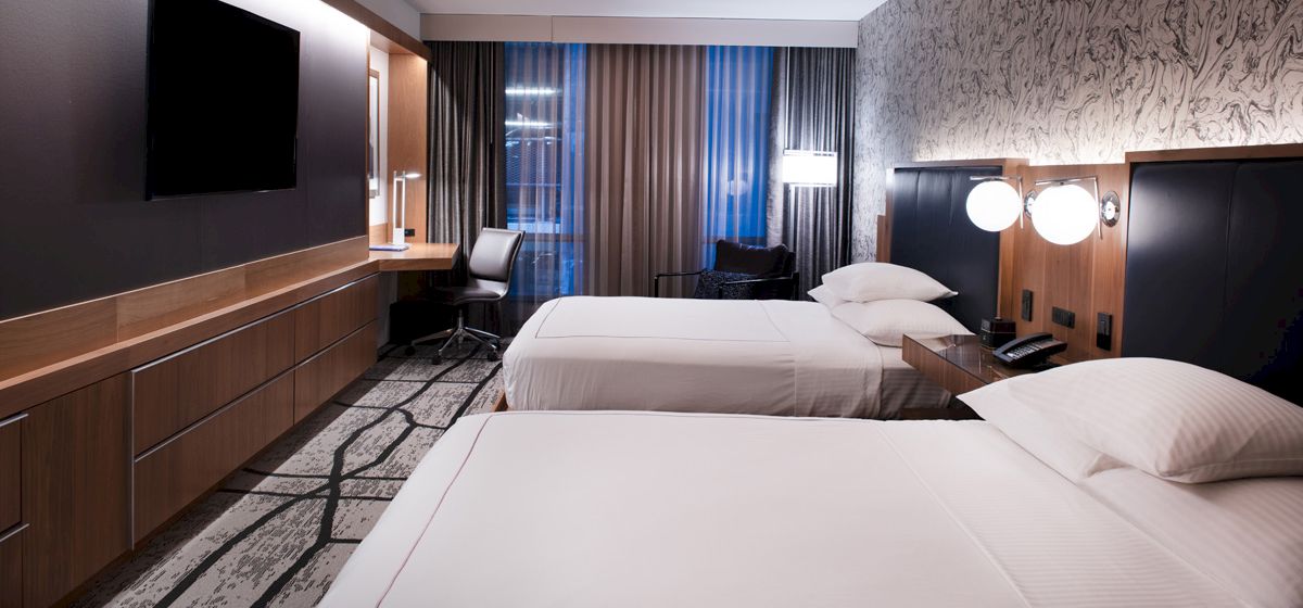 The image shows a modern hotel room with two beds, a TV, a desk with a chair, and minimalist decor, featuring neutral tones and a patterned carpet.