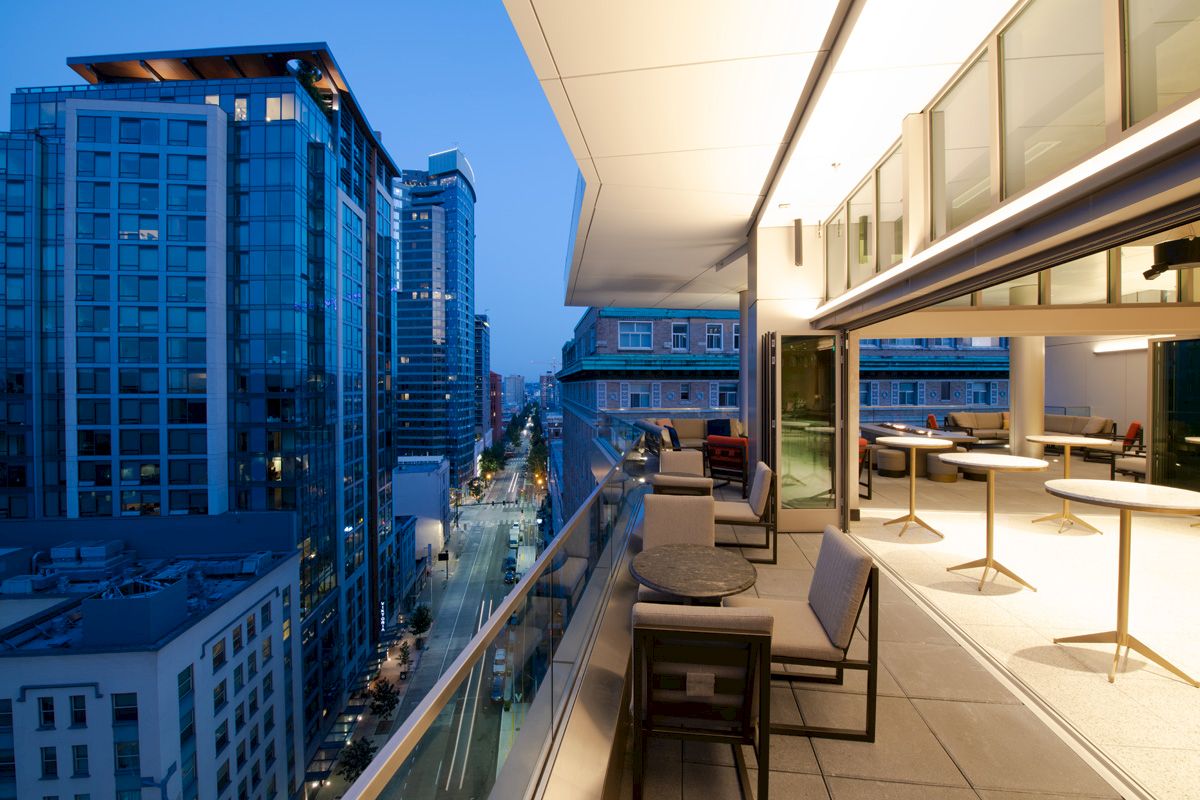 A modern urban balcony at twilight with a cityscape view, featuring outdoor seating and tables, overlooking street and high-rise buildings.