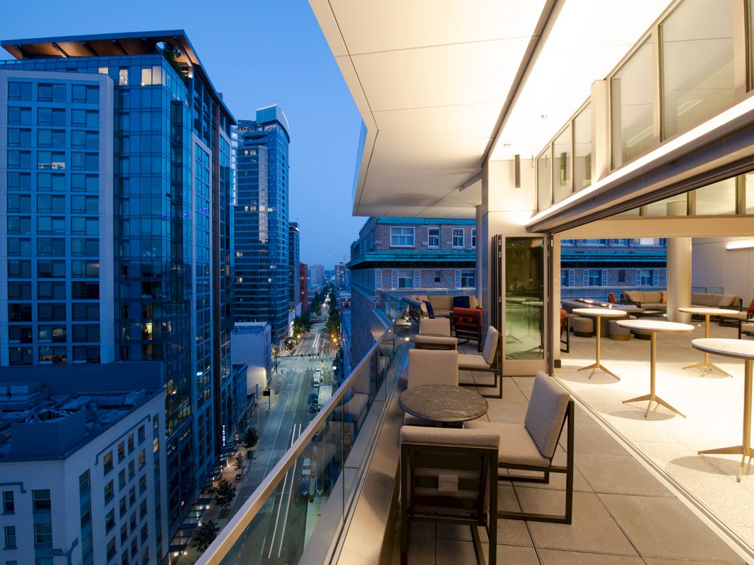A modern urban balcony at twilight with a cityscape view, featuring outdoor seating and tables, overlooking street and high-rise buildings.