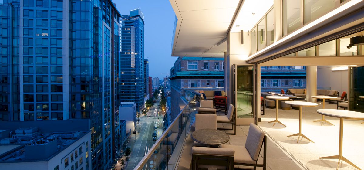 A modern urban balcony at twilight with a cityscape view, featuring outdoor seating and tables, overlooking street and high-rise buildings.
